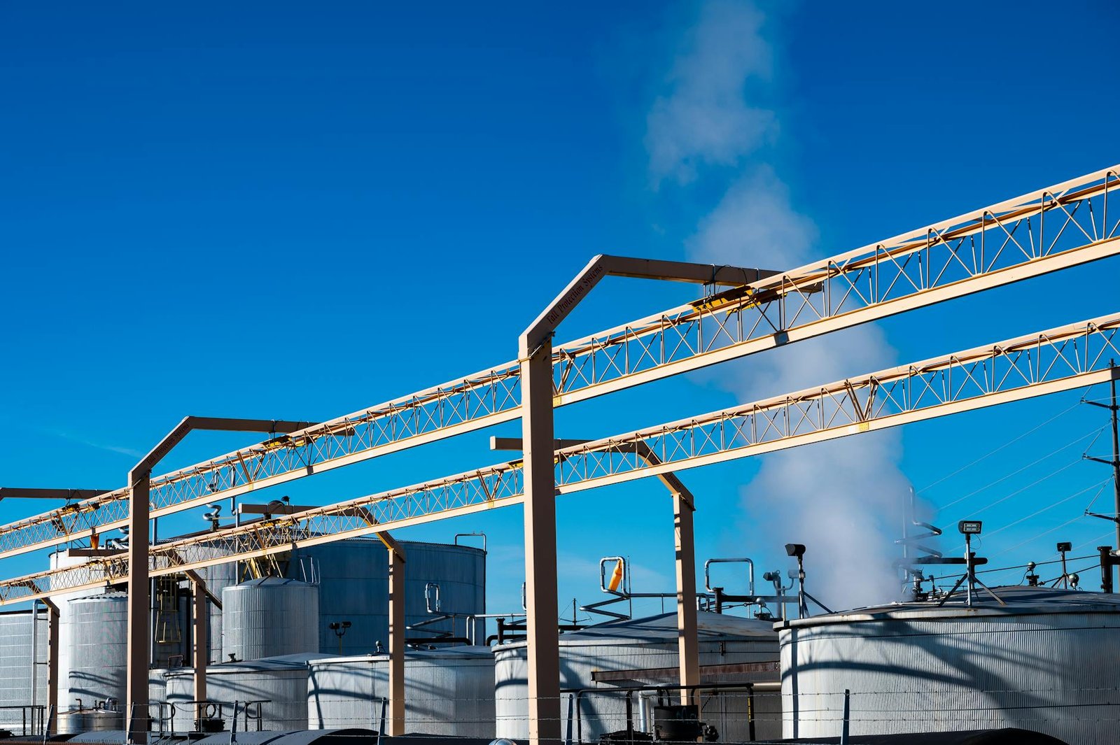 Wide view of an industrial plant with smoke rising from storage tanks against a clear blue sky.