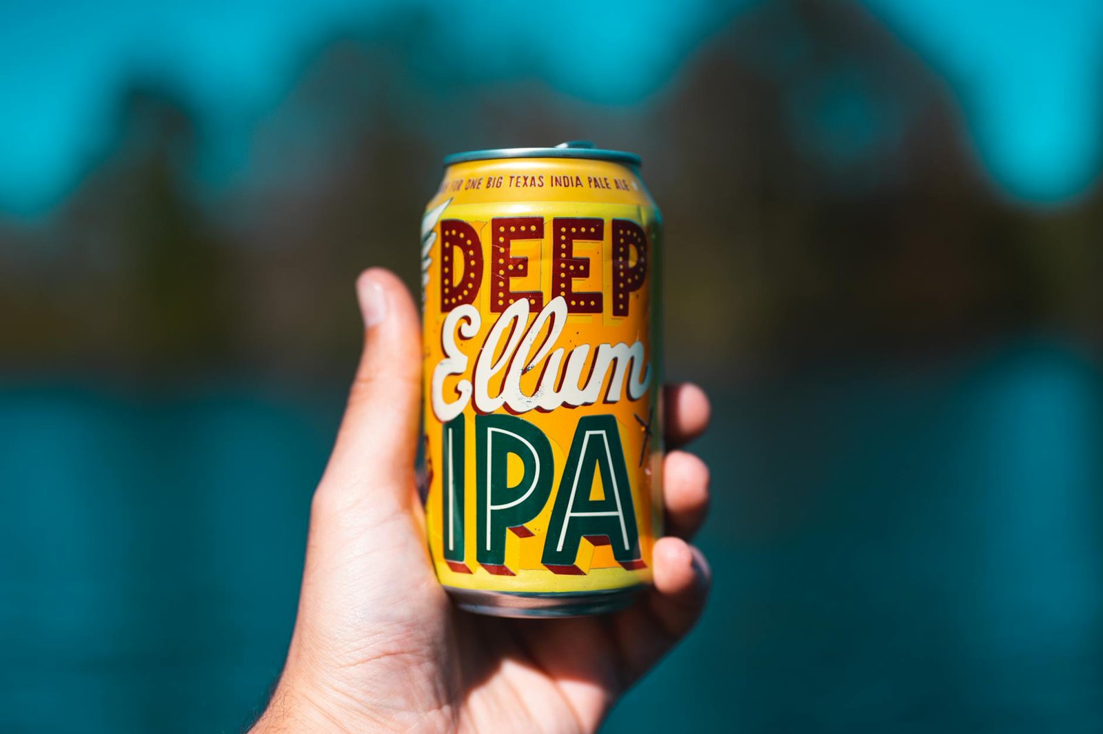 Close-up of a hand holding a Deep Ellum IPA can with a blurred outdoor background.